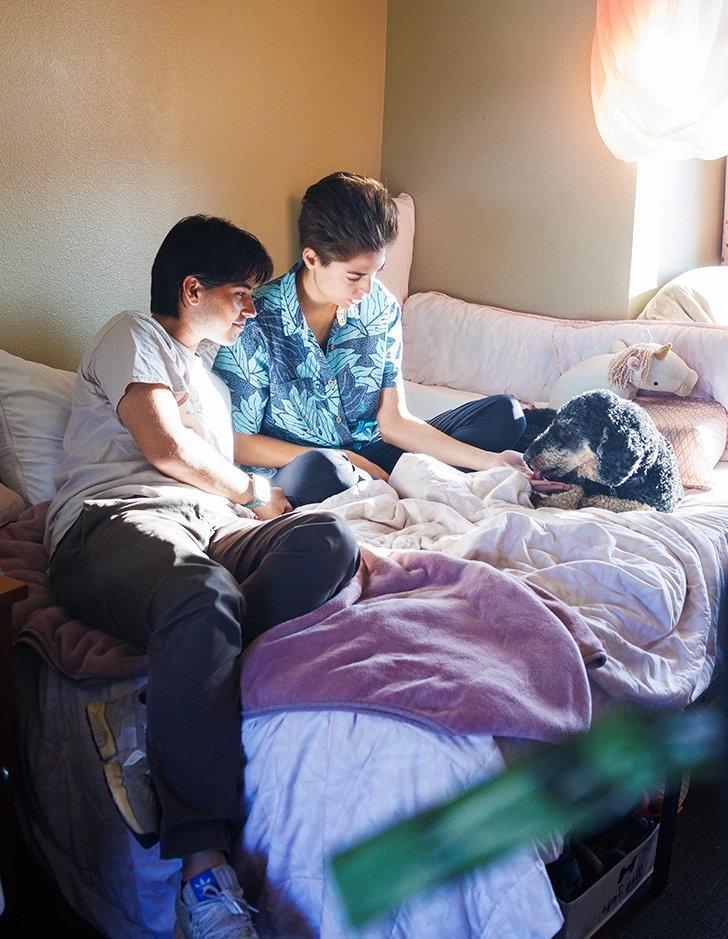 two students sit on a dorm bed and pet a dog